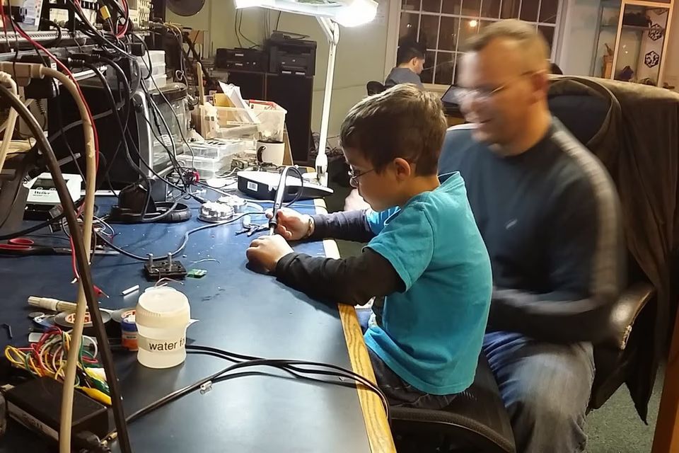 Young boy wearing glasses focused on soldering a circuit board at a well-equipped electronics workstation with an adult mentor beside him
