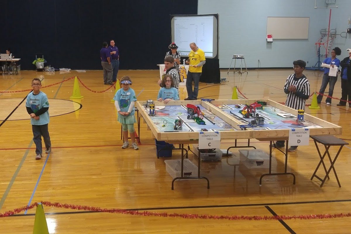 FIRST LEGO League competition with kids in team shirts operating their robots on the competition table while referees watch