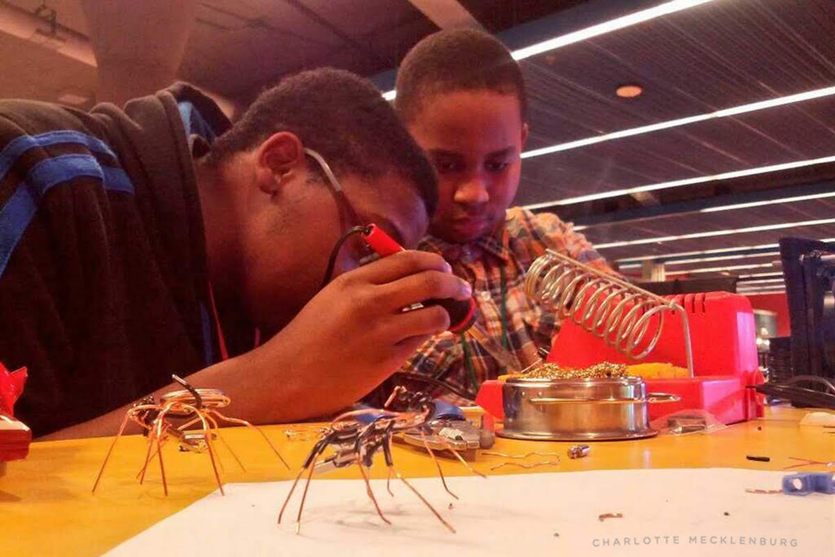Two boys working together at a soldering station, carefully assembling electronic bug bot sculptures with copper wire legs
