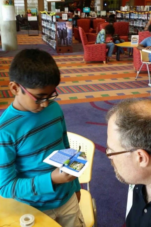 Boy at Imaginon library showing off his motorized paper airplane creation to an instructor, demonstrating how the propeller attachment works