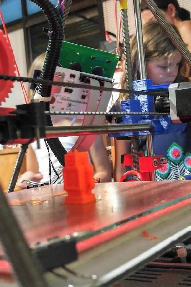 Young child watching a Prusa 3D printer in action, observing an orange object being printed layer by layer